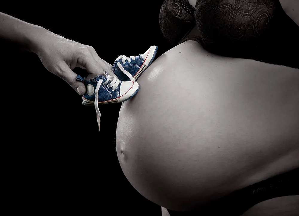 Schwarz-Weiß-Foto eines Schwangerschaftsbauchs mit kleinen blauen Babyschuhen auf dem Bauch – Schwangerschafts- und Babyfotografie im Fotostudio Foto Ape in Rostock